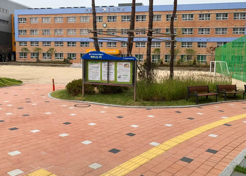 강덕초등학교 운동장 앞 통로. 적갈색 블록 바탕에 짙은 회색·흰색 블록이 점점이 섞여 있고 노란 점자블록 선이 지나며 뒤로 교사동이 보인다.