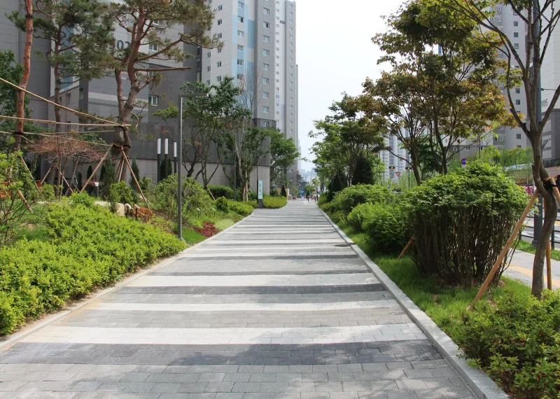 천안 불당 푸르지오 녹지 보행로. 회색 블록 길에 짙은 회색·흰색 가로 띠가 반복되며 관목 사이로 이어진다.