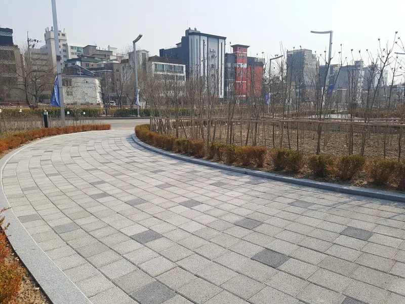 수원보훈원 곡선 산책로를 반대 방향에서 본 모습. 회색 블록 길 가장자리를 화강석 경계석과 붉은빛 관목이 따라간다.