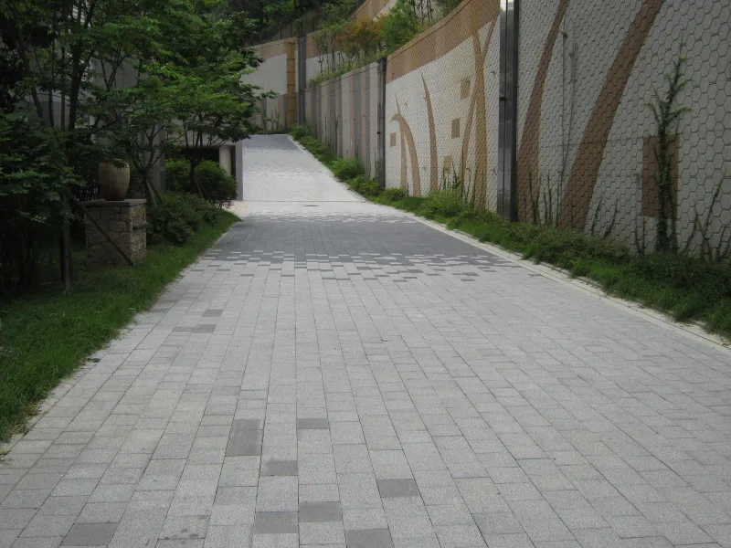 현대 엠코타운 옹벽 옆 경사 통로. 연회색 블록 길이 장식 타일 벽과 화단 사이로 비탈을 따라 올라간다.