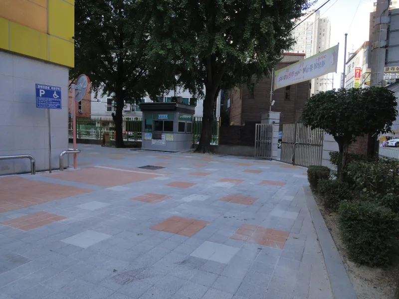 중계초등학교 후문 쪽 진입로. 회색 블록 바닥에 적갈색·흰 블록 사각 무늬가 흩어진 넓은 길이 교문과 경비실 쪽으로 이어진다.