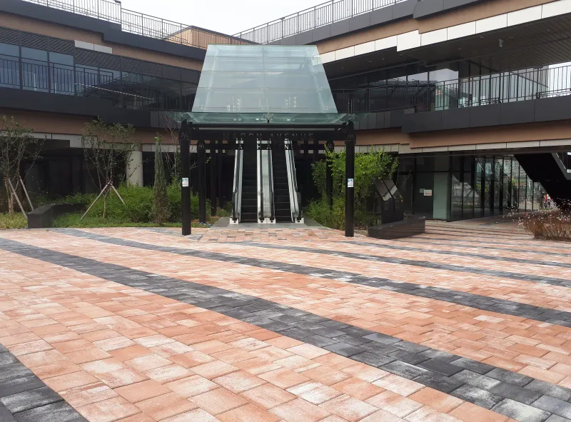 마치에비뉴 2차 에스컬레이터 입구 앞. 유리 지붕 출입구까지 적갈색 블록과 짙은 회색 띠가 비스듬히 깔려 있고 양옆으로 나무 화단이 있다.