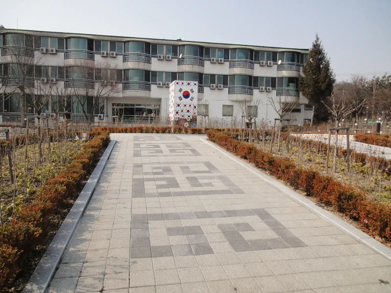 수원보훈원 건물 쪽 보행로. 밝은 회색 블록 바닥 가운데 짙은 회색 블록으로 만든 사각 번개무늬가 반복되고 양옆에 관목 화단이 있다.