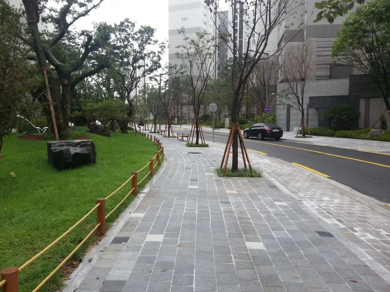 아현동 현장의 공원 옆 보도. 회색 톤이 섞인 직사각형 블록이 잔디 화단의 밧줄 울타리를 따라 완만하게 휘어 이어지고, 가로수 너머로 아파트 단지가 보인다.