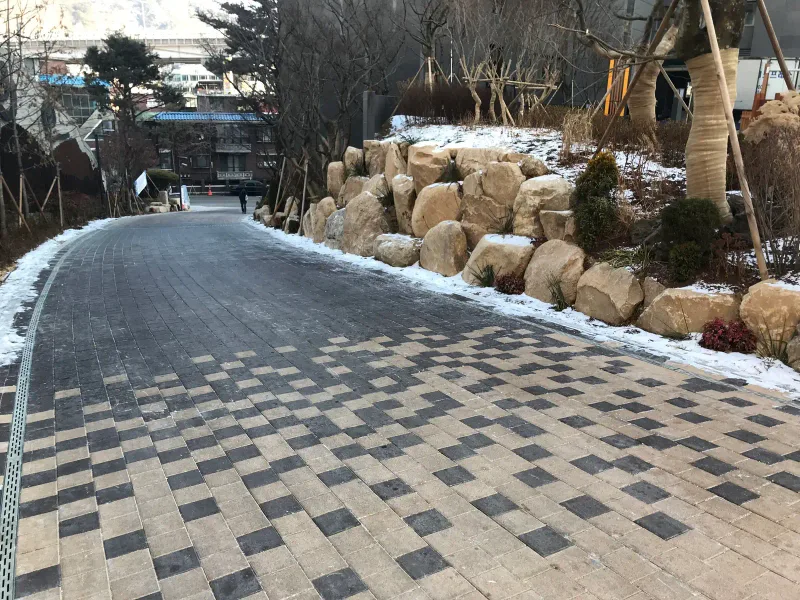 북한산 더샵 단지 내 진입로. 짙은 회색에서 베이지로 번져 가는 바둑판 무늬 블록 길이 자연석 조경 옆으로 오르막을 이룬다.
