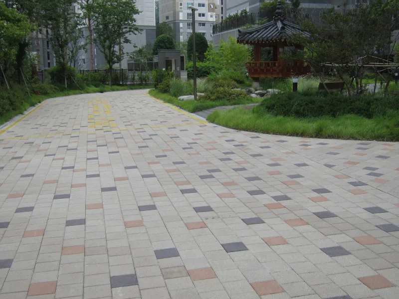 현대 엠코타운 단지 내 정자 앞 통로. 베이지색 사각 블록 사이에 짙은 회색·적갈색 블록이 점점이 박혀 있고 곡선 화단 너머로 전통 정자가 보인다.