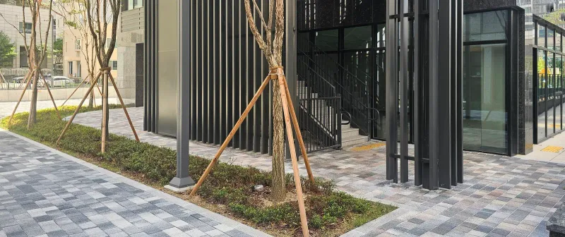 대구 달서 푸르지오 시그니처 건물 옆 화단 길. 회색·분홍빛이 섞인 블록 보행로가 나무를 심은 화단을 따라 계단실 출입구 앞까지 이어진다.