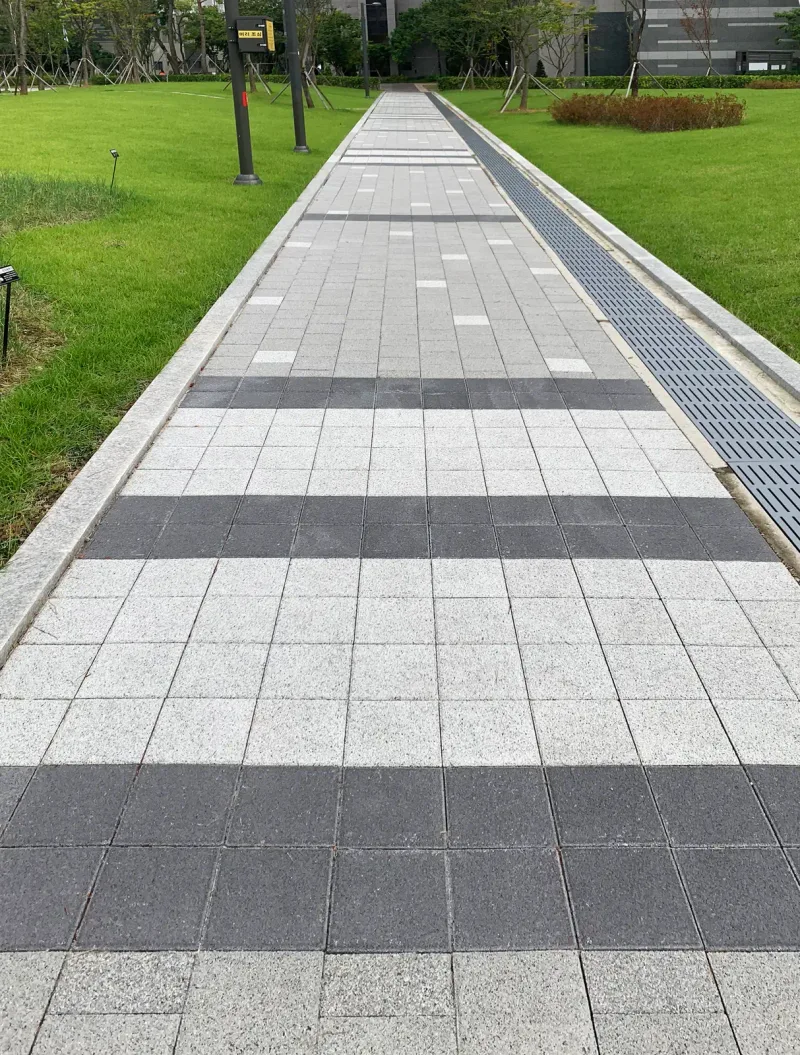 고덕근린공원 잔디밭 사이 곧은 산책로. 배수 트렌치를 낀 회색 블록 길에 짙은 회색 띠가 규칙적으로 반복되며 멀리 이어진다.