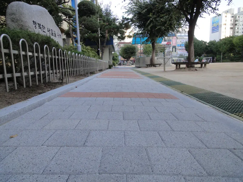 중계초등학교 교명석 옆 통로. 밝은 회색 블록 길에 적갈색 블록 띠가 지나고 운동장 쪽으로 배수 그레이팅이 나란히 깔려 있다.