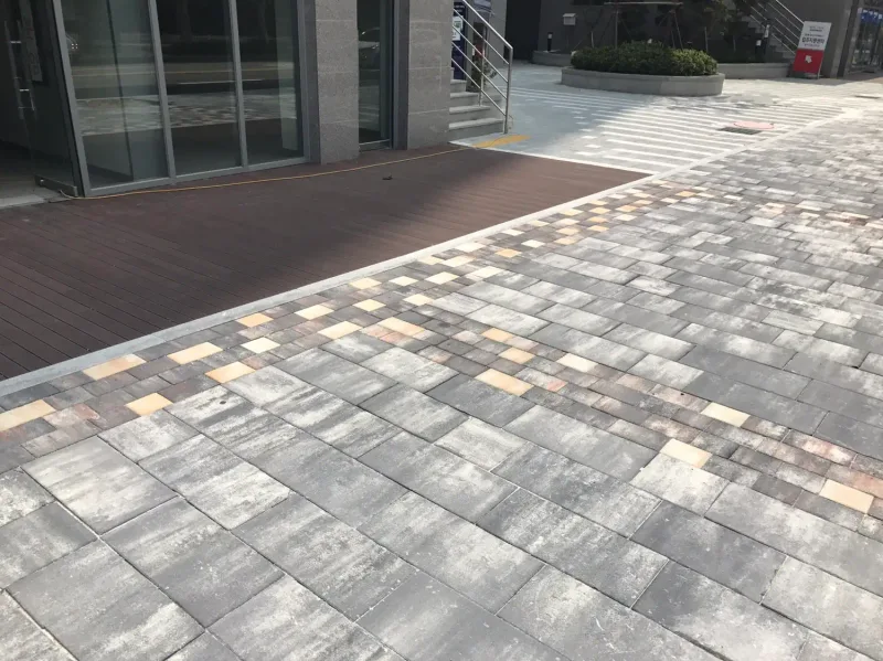 석촌호수 카페거리 건물 출입구 앞. 목재 데크 가장자리를 따라 베이지·분홍빛 블록 띠가 들어가고 그 바깥으로 회색 섞임 블록이 이어진다.