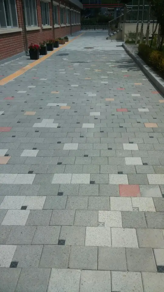 위례 한빛초등학교 교사동 옆 긴 통로. 밝은 회색 블록 바탕에 짙은 회색·분홍·베이지 블록이 흩어져 깔려 있고 가장자리에 화단이 있다.