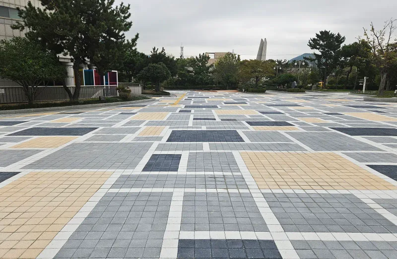 을숙도 들락날락 광장 입구 방향. 베이지·회색·짙은 회색 블록 구획이 흰 띠 사이에 엇갈려 배치되어 있고 뒤로 소나무 숲이 이어진다.