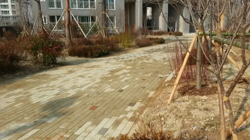 전주 우미린 아파트 단지 조경 산책로. 베이지·연노랑 블록에 회색 블록이 섞여 깔린 길이 어린 나무와 관목 화단 사이로 이어지고 뒤로 아파트 동이 보인다.