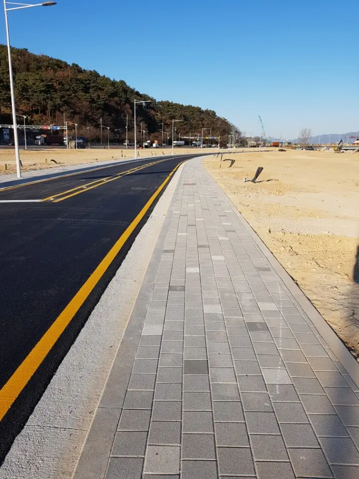 부산 범방동 새 도로변 보도. 회색 계열 직사각형 블록이 길게 깔린 인도가 노란 중앙선 도로를 따라 뻗어 있고 뒤로 산과 빈 부지가 보인다.