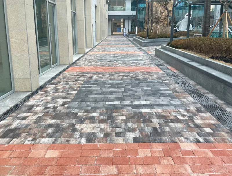 한강미사 현대지식산업센터 2차 건물과 화단 사이 긴 통로. 회색·갈색 섞임 블록이 깔리고 적갈색 블록 띠가 일정 간격으로 가로지른다.