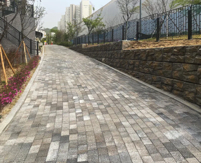 위례 자이 더 시티 단지 경사 산책로. 회색·베이지가 섞인 직사각형 블록이 오르막길을 따라 길게 깔려 있고 오른쪽에 석축과 난간, 멀리 아파트가 보인다.