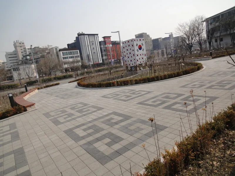 수원보훈원 광장 곡선부. 짙은 회색 번개무늬가 박힌 회색 블록 바닥이 원형 화단을 감싸 돌고 뒤로 도심 건물이 보인다.