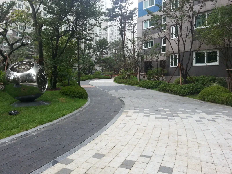 아현동 현장 아파트 단지 안 곡선 산책로. 짙은 회색 블록 테두리와 밝은 회색 블록 바닥이 부드럽게 휘어지고, 옆 잔디밭에 금속 구 조형물이 놓여 있다.