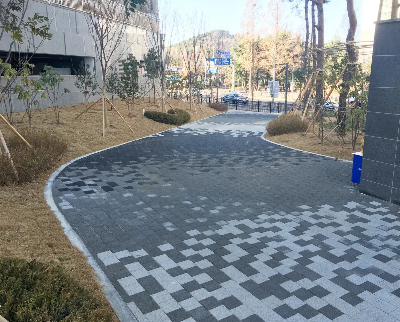 창원 용호 더샵 동 옆 곡선 보행로. 짙은 회색 블록 길 위로 밝은 회색 블록이 계단식 모자이크로 번져 나가며 도로 쪽으로 휘어 간다.