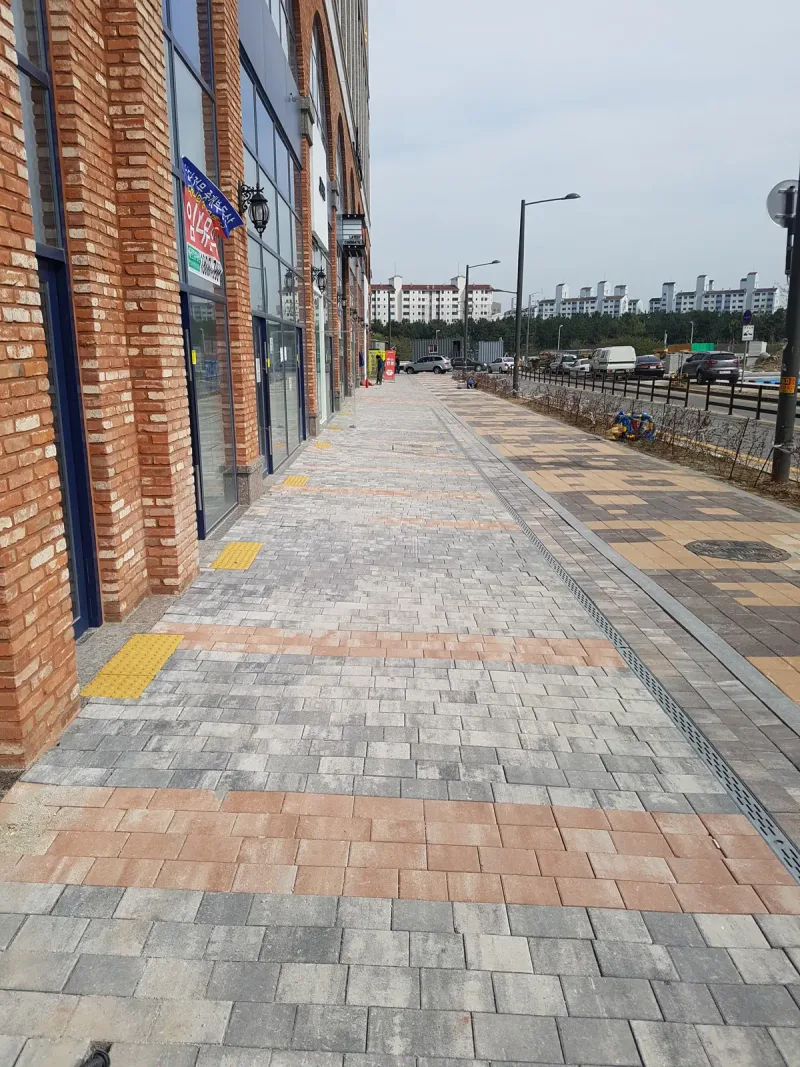 베니스스퀘어 도로 쪽 상가 전면 보도. 회색과 분홍빛 블록이 섞인 길에 베이지 가로 띠와 노란 점자블록이 들어가 있다.