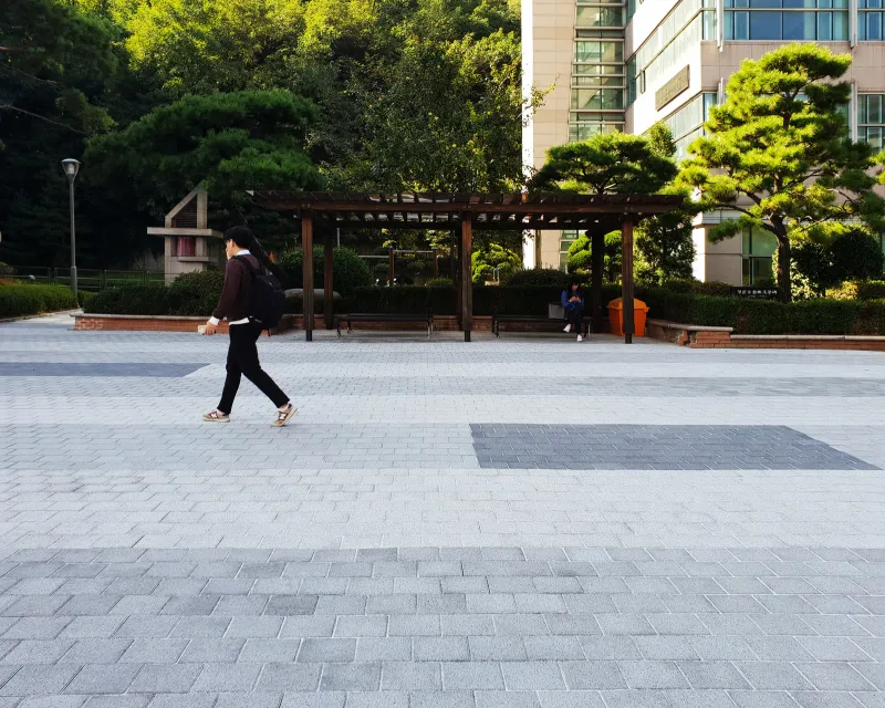 중앙대학교 쉼터 앞 광장. 밝은 회색 블록 바닥에 짙은 회색 블록 띠가 사각으로 들어가 있고 뒤로 목재 퍼걸러와 소나무가 보인다.