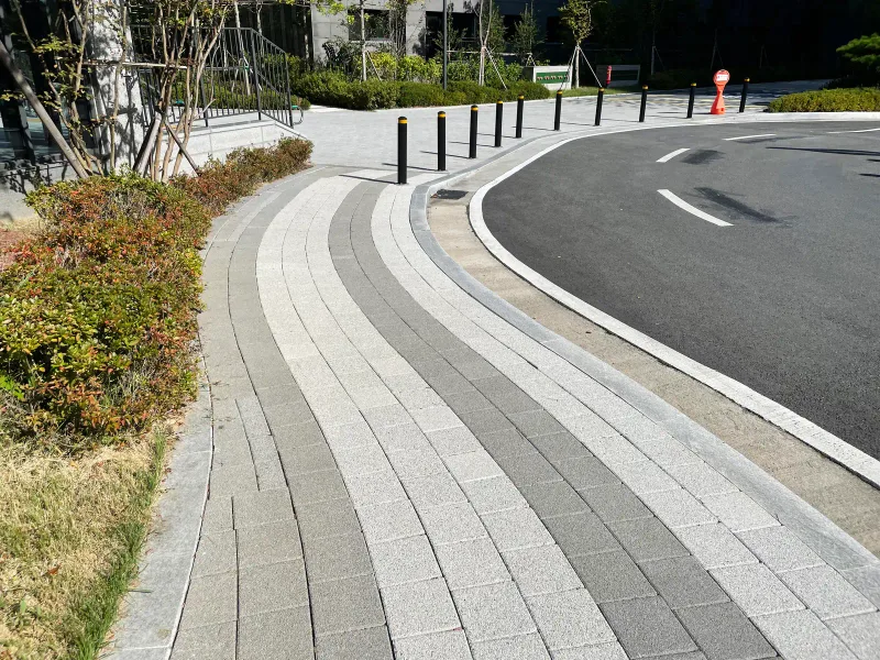 e편한세상 지축 센텀가든 차도 옆 곡선 보도. 흰색·연회색·짙은 회색 블록이 긴 줄무늬로 도로 곡선을 따라 휘어지고 검은 볼라드가 줄지어 있다.