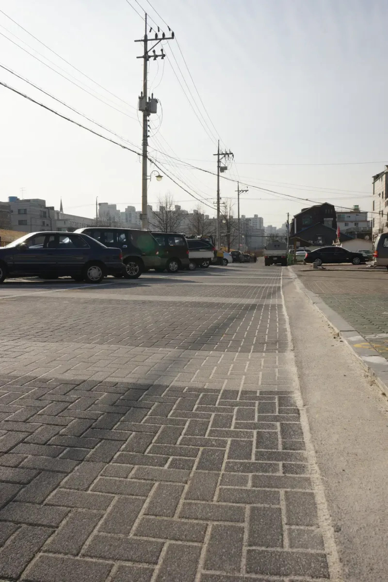 풍납동 도로변 보도 세로 사진. 회색 블록이 헤링본으로 깔린 보도가 경계석을 따라 곧게 뻗고 멀리 주차 차량과 전신주가 보인다.