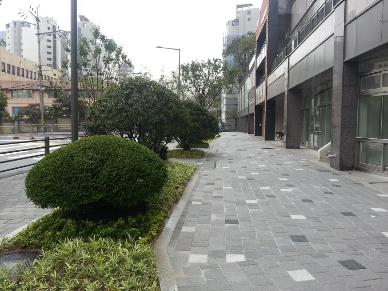 아현동 현장 상가 건물 앞 보도. 밝은 회색 블록 사이에 짙은 블록이 드문드문 섞인 넓은 보도가 둥근 관목 화단 옆으로 곧게 뻗어 있다.