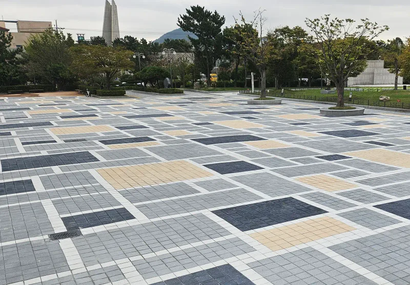 을숙도 들락날락 광장 전경. 회색 바탕에 짙은 회색·베이지색 사각 구획을 흰 띠로 나눠 깔았고 뒤로 원형 화단과 나무가 보인다.
