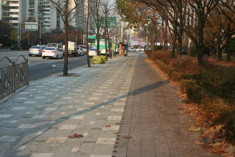풍납동 버스정류장 앞 보도. 낙엽이 흩어진 밝은 회색·회색 섞임 블록 구간과 화단 쪽 회색 블록 구간이 나란히 이어지고 옆으로 차도가 지난다.