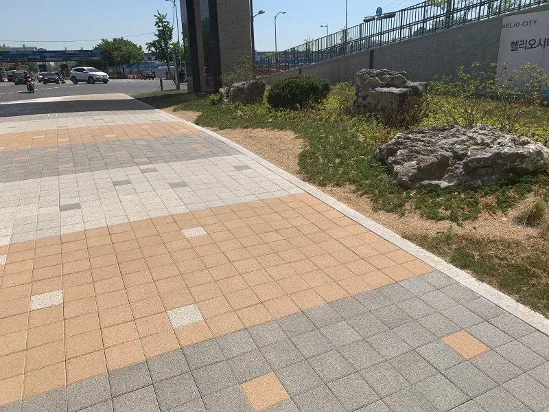 송파 헬리오시티 조경석 옆 보도. 주황빛 블록 띠와 짙은 회색 블록 띠가 번갈아 놓이고 가장자리에 자연석 화단이 있다.
