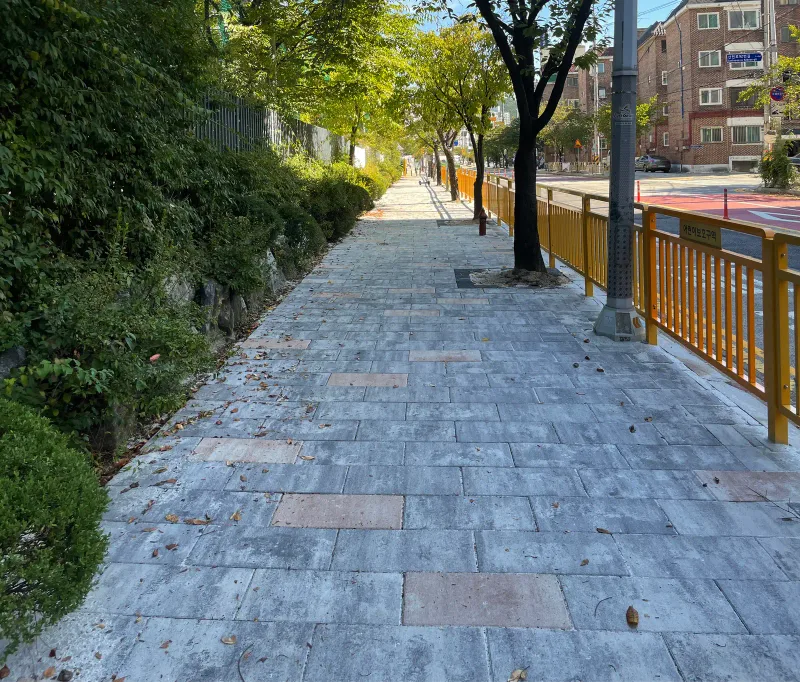 고양 현산초등학교 일원 보도 원경. 회색 블렌딩 판형 블록 길이 가로수와 노란 펜스를 따라 곧게 뻗고 건너편에 주택가가 보인다.