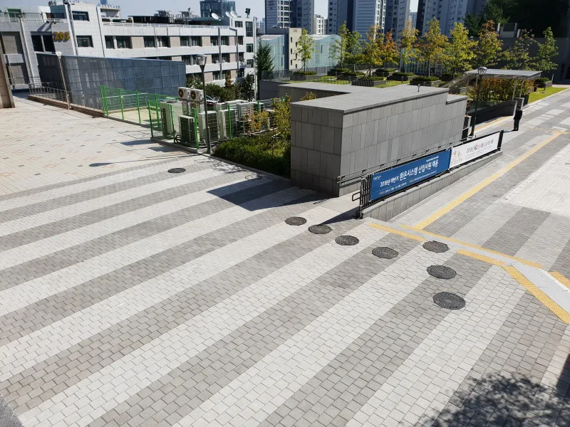 중앙대학교 캠퍼스 광장. 연회색·짙은 회색·흰색 블록이 넓은 줄무늬로 깔린 바닥 위에 환기 구조물과 조경 화단이 놓여 있다.