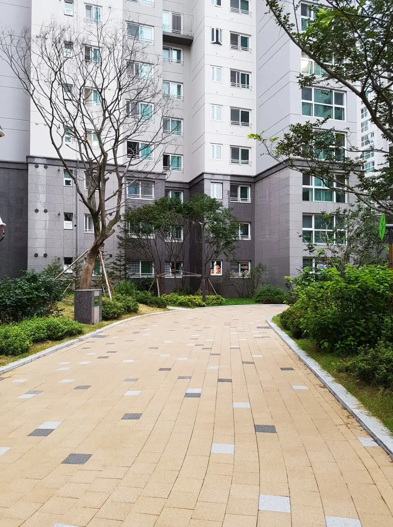 신길 래미안 아파트 동 앞 보행로 세로 사진. 베이지 블록 길이 관목 화단 사이로 뻗어 동 입구 쪽 나무 아래로 이어진다.