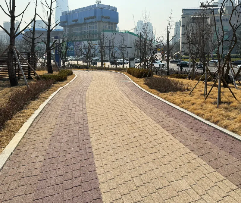 올림픽회관 녹지 사이 곡선 산책로. 베이지 블록 바탕에 적갈색 블록 띠 두 줄이 길을 따라 부드럽게 휘어진다.