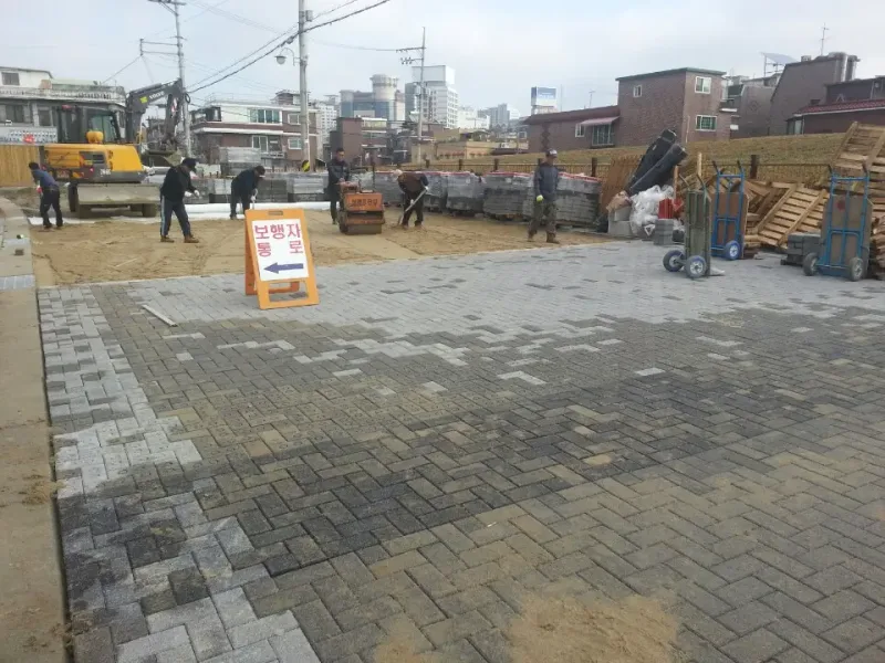 송파 저류기층 시범구간, 시공 중. 짙은 회색과 밝은 회색 블록을 헤링본 무늬로 깔아 나가는 도로 앞에 보행자 통로 안내판이 서 있고, 뒤로 다짐 장비와 블록 더미가 보인다.
