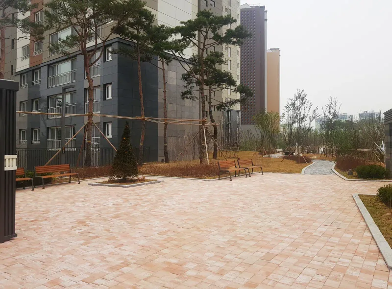 동탄2 아이파크 아파트 동 앞 마당. 연분홍 계열 블록 바닥 가장자리에 소나무와 벤치가 있고 안쪽으로 회색 길이 이어진다.