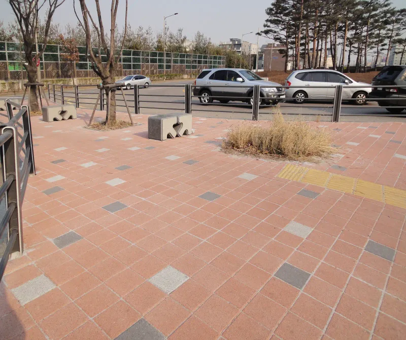 수원 세류동 보도 쉼터 공간. 적색 정사각 블록 바탕에 회색·흰색 블록이 흩어져 있고 돌 의자와 노란 점자블록, 가로수가 놓여 있다.
