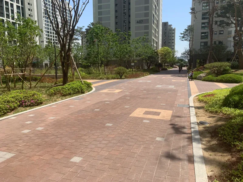 송파 헬리오시티 곡선 산책로. 적갈색 블록 바닥에 주황색 네모 무늬가 들어가고 둘레에 관목 화단이 이어진다.
