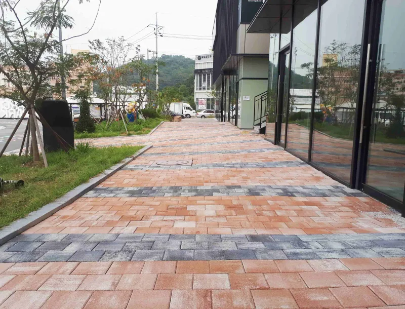 마치에비뉴 2차 상가 유리벽을 따라 난 보도. 주황빛 블렌딩 블록에 회색 띠가 가로로 들어가고 왼쪽에 잔디 화단과 가로수가 있다.