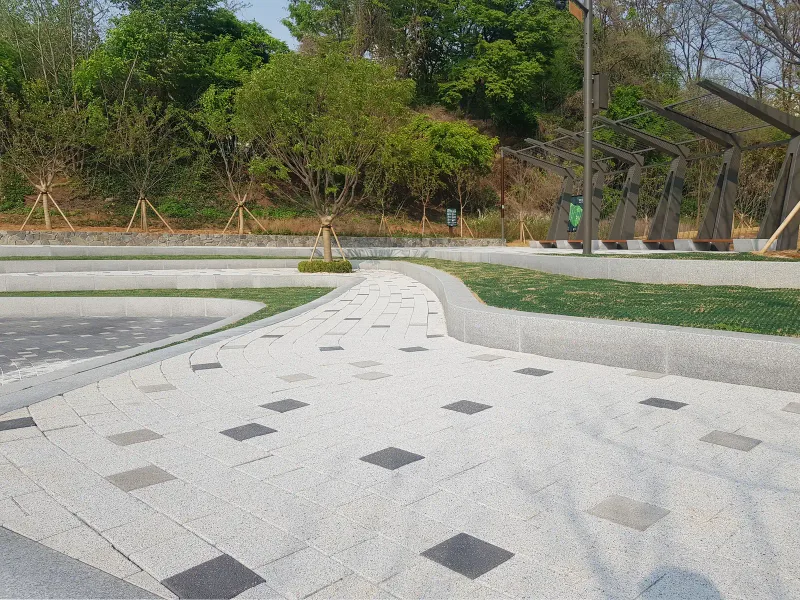 개포 래미안 조경 공간. 흰빛이 도는 밝은 블록에 짙은 회색 사각 블록을 흩뿌리듯 섞었고 곡선 경계석 너머로 잔디와 숲이 있다.