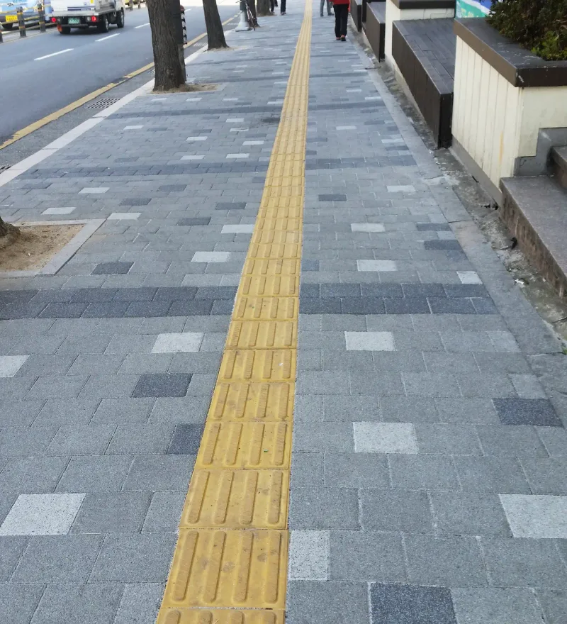 소방안전본부 일대 보도 근접(세로 사진). 회색 블록 사이에 흰색·짙은 회색 블록이 흩어져 있고 노란 점자블록 줄이 곧게 지나간다.