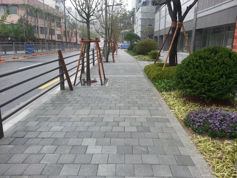 아현동 현장 보도를 따라 본 모습. 회색 블록 보도 한쪽에 지지대를 세운 가로수와 난간, 다른 쪽에 보라색 꽃이 핀 화단이 나란하다.