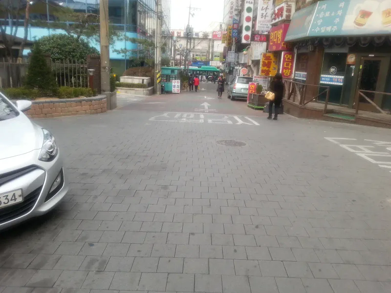 부천 소사 테마거리의 다른 골목. 회색 직사각형 블록이 엇갈려 깔린 바닥에 흰색 노면 글자와 화살표, 맨홀이 있고 길가로 상가와 화단이 이어진다.