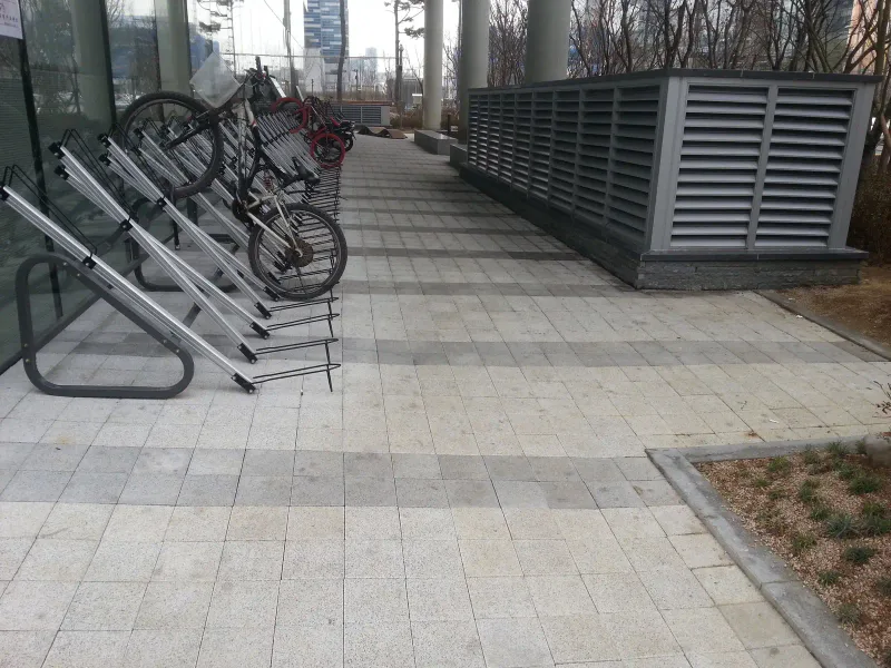 판교 SK 사옥 옆 통로. 밝은 회색 블록 바닥을 따라 자전거 거치대와 회색 환기 구조물이 나란히 있고, 짙은 회색 블록 띠가 경계를 이룬다.