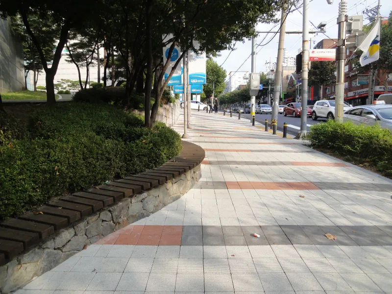 숭실대학교 중문 앞 쉼터 구간. 둥글게 휜 나무 좌석 화단 옆으로 적갈색·회색 줄무늬 블록 보도가 도로 쪽으로 이어진다.