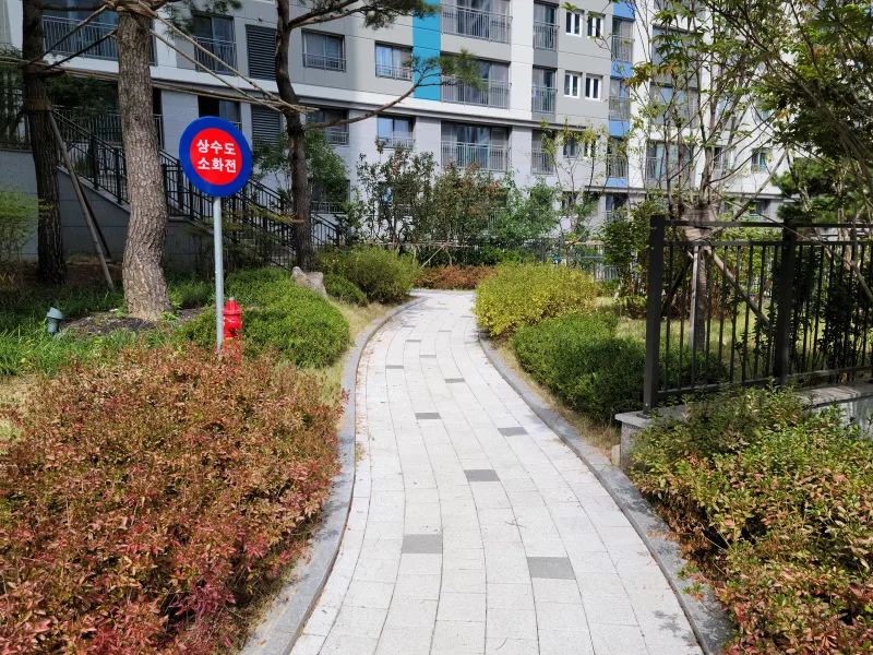 더샵 동천 이스트포레 단지 안 곡선 산책로. 밝은 회색 블록 길이 붉은 관목과 초록 화단 사이로 부드럽게 굽어 나간다.