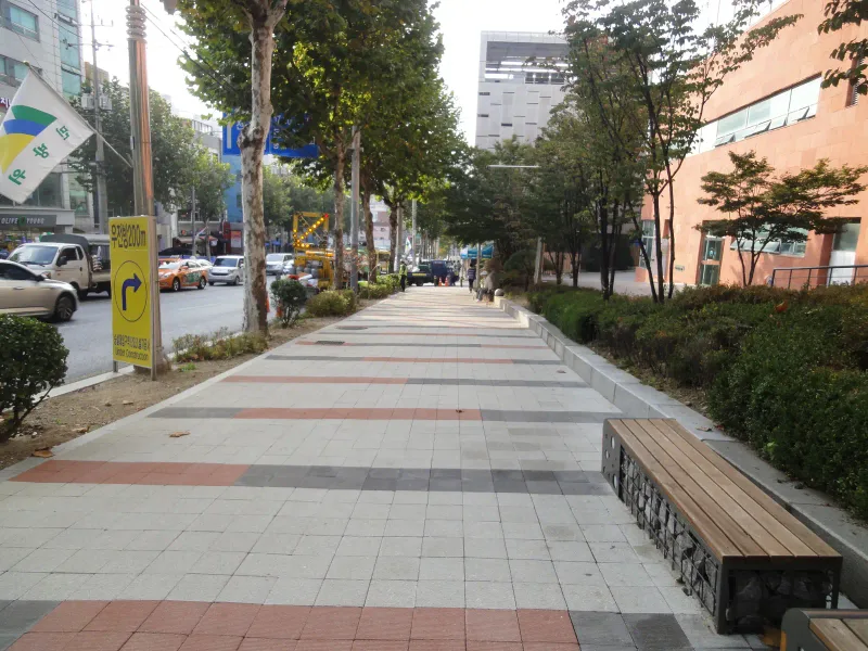 숭실대학교 중문 앞 보도 벤치. 돌망태 받침 나무 벤치가 놓인 연회색 블록 보도에 적갈색·짙은 회색 줄이 가로지르고 뒤로 가로수 길이 뻗는다.