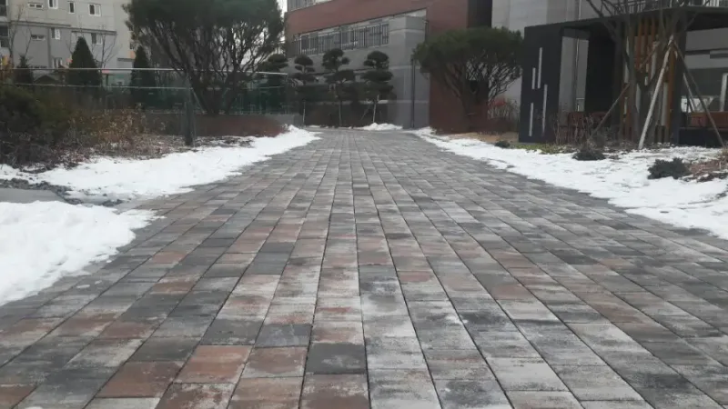 에일린의 뜰 단지 안 곧은 길. 짙은 회색·연회색·적갈색이 섞인 직사각형 블록이 눈 쌓인 화단 사이로 곧게 이어진다.