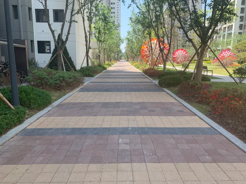 송파 헬리오시티 단지 내 산책로. 연적갈·베이지·짙은 회색 블록이 가로 띠를 이루며 길게 깔려 있고 옆으로 꽃 조형물과 아파트가 보인다.